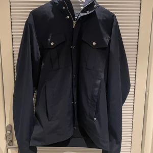 J.lindeberg Field jacket - Säljer en snygg mörkblå jacka från J.Lindeberg i mycket bra skick, använd kanske 2 max 3 gånger Jackan har en dragkedja och knappar framtill, samt praktiska fickor med lock. Perfekt för höst och vår, med en stilren design som passar både till vardags och lite finare tillfällen. Kan givetsvis skicka mer bilder bara att höra av er. Den är i storlek L men jag använder det som bär storlek M. Tack på förhand! (Inga defekter)