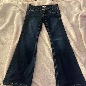 Mörkblå bootcut jeans - Säljer ett par snygga mörkblå bootcut jeans i bra skick. De har vita kontrastsömmar och klassiska femficksdetaljer med knappar på bakfickorna. Perfekta för en avslappnad men stilren look. Passar både till vardags och fest!