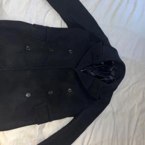 Svart kappa från Mango - Säljer en stilren svart kappa i mycket bra skick. Den har en klassisk dubbelknäppning med snygga knappar och en elegant krage. Perfekt för höst och vinter, och funkar både till vardags och fest. Passar bra över en tröja eller skjorta. Hör av dig vid intresse!