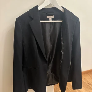 Svart kavaj - Snygg svart kavaj i klassisk stil. Perfekt för både formella och semi-formella tillfällen. Den har en elegant passform och är tillverkad i ett material som ger en stilren look. Ett måste i garderoben för alla som vill ha en tidlös stil.