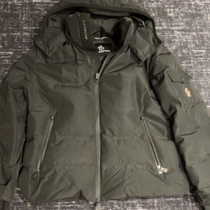 Svart vinterjacka från Moncler  - Säljer en snygg svart dunjacka från Moncler i mycket bra skick. Jackan har en dragkedja framtill och praktiska fickor med dragkedjor. Perfekt för kalla vinterdagar med sin varma och stilrena design. Den har en avtagbar huva och Moncler-logga på ärmen. Passar både till vardags och fest!