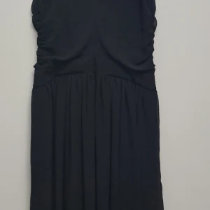 Svart klänning - Elegant svart klänning med draperad design och smickrande passform. Perfekt för festliga tillfällen. Materialet ser ut att vara mjukt och bekvämt, vilket ger en fin fall. Klänningen har en klassisk stil som aldrig går ur modet.