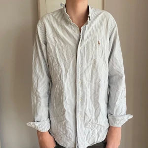 Randig skjorta från Ralph Lauren - Säljer en stilren och klassisk randig skjorta från Ralph Lauren i storlek M. Skjortan är i bomull och har en snygg broderad logga på bröstet. Perfekt för både vardag och fest! Den har långa ärmar och knappar hela vägen ner. Passformen är custom fit vilket ger en snygg siluett. Lite skrynklig på bilden eftersom inte strykt. Modellen är 176cm