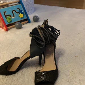 Svarta klackskor med snörning - Snygga svarta klackskor med öppen tå och elegant snörning runt ankeln. Perfekta för festliga tillfällen. Skorna har en texturerad yta och en bekväm klackhöjd. De är i bra skick och redo för nästa ägare!