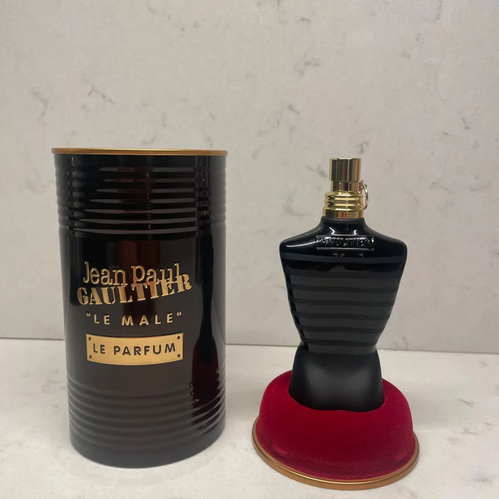 Säljer en elegant parfym från Jean Paul Gaultier, 'Le Male Le Parfum'. Flaskan är formad som en svart torso och står på en röd bas. Den kommer i en stilren svart metallförpackning med guldtext. Perfekt för den som vill ha en sofistikerad och maskulin doft.. Parfym.