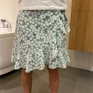Blommig omlottkjol i grönt och vitt - Superfin blommig omlottkjol i grönt och vitt med volangdetaljer. Perfekt för sommardagar eller en mysig vårutflykt. Kjolen har en knytning i sidan som ger en söt touch. Materialet känns lätt och luftigt, vilket gör den idealisk för varmare väder. Säljes då den inte kommer till användning längre!