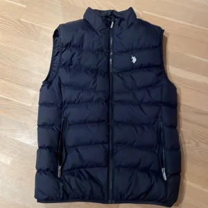 Svart dunväst från U.S. Polo Assn. - Säljer en snygg svart dunväst från U.S. Polo Assn. i storlek S. Västen har en dragkedja framtill och är quiltad för extra värme. Perfekt för höst och vår! Den har en broderad logga på bröstet och två praktiska fickor med dragkedja.