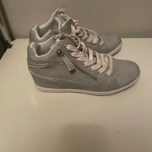 Grå sneakers med dragkedja - Säljer ett par snygga grå sneakers i mycket bra skick. De har både snörning och en praktisk dragkedja på sidan för enkel på- och avtagning. Perfekta för vardagsbruk och passar till de flesta outfits. Skorna har en stilren design med en bekväm passform.