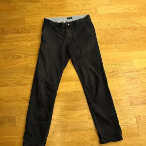 Svarta chinos från GANT - Säljer ett par stilrena svarta/mörk blåa chinos från GANT. De är i bra skick och perfekta för både vardag och fest. Byxorna har en klassisk passform med knappar och dragkedja. Insidan har ett snyggt randigt mönster. Passar perfekt till höst och vår! 8/10 skick!