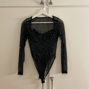 Svart glittrig bodysuit - Snygg svart bodysuit med glittriga detaljer. Den har långa ärmar och en åtsittande passform, perfekt för festliga tillfällen. Materialet är genomskinligt på ärmarna och över axlarna vilket ger en elegant look.