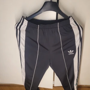 Svarta träningsbyxor från Adidas - Snygga svarta träningsbyxor från Adidas med vita detaljer och klassiska ränder längs sidorna. Byxorna har en elastisk midja och broderade blommor på ena benet för en unik touch. Perfekta för både träning och vardagsbruk!