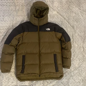 Olivgrön dunjacka från The North Face - Säljer en olivgrön dunjacka från The North Face i mycket bra skick. Jackan har ett svart axelparti och huva, perfekt för kalla vinterdagar. Den är långärmad med dragkedja och två praktiska fickor framtill och en bröstficka på insidan. Supervarm och stilren för vintern! ❄️