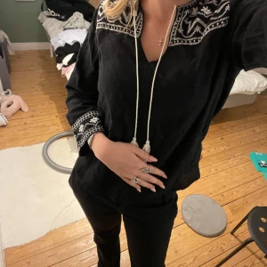 Svart blus med broderi - Säljer en svart blus med vackra vita broderier vid halsen och ärmsluten. Blusen har en avslappnad passform och långa ärmar. Perfekt för både vardag och fest! 🖤