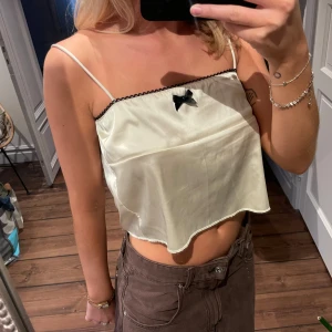 Vit satinlinne med rosett - Säljer ett supergulligt vitt satinlinne med smala axelband och en söt svart rosett framtill. Toppen har en öppen rygg med knytband, vilket ger en extra touch. Perfekt för sommardagar eller en utekväll. Passar till både jeans och kjol!