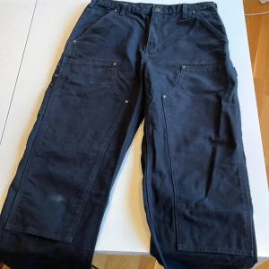 Svarta arbetsbyxor från Carhartt - Säljer ett par robusta svarta arbetsbyxor från Carhartt. Byxorna har flera praktiska fickor och förstärkta detaljer med nitar. Perfekta för arbete eller fritid där slitstyrka behövs. De har en klassisk passform och är gjorda i slitstarkt material. lite slitage på höger knä.