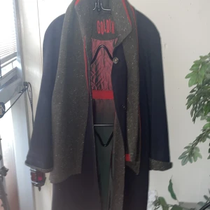 Mörk kappa från Goldix - Säljer en elegant mörk kappa från Goldix med tillhörande schal.Den har en quiltad röd insida och en stilren design med knappar framtill. Perfekt för höst och vinter, med en klassisk look som passar både till vardags och fest.