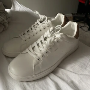 Vita sneakers från ONLY - Säljer ett par vita sneakers från ONLY i nytt skick. De har snygga guldfärgade detaljer på hälen som ger en glittrig touch. Skorna har klassisk snörning och är perfekta för både vardag och fest. Sulan är vit och har bra grepp. Passar perfekt till vår och sommar! Köptes på Zalando för 479, säljer dem helt nya oanvänd för 250, lådan medföljer ej.