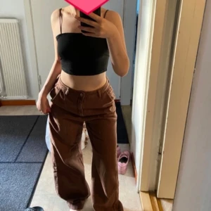 Bruna byxor - Säljer ett par snygga bruna byxor i loose fit. De är högmidjade och har en klassisk design med knappar och dragkedja. Perfekta för en avslappnad stil och kan matchas med en crop top för en trendig look.