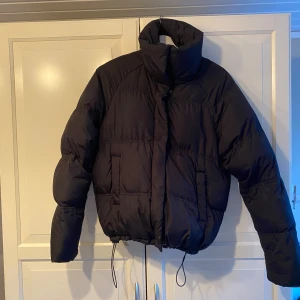 Svart pufferjacka - Säljer en svart pufferjacka i mycket bra skick. Den är perfekt för kalla dagar med sin tjocka vaddering och höga krage. Jackan har långa ärmar och dragsko i nederkant för extra värme. Passar perfekt till vinterns kyliga väder!