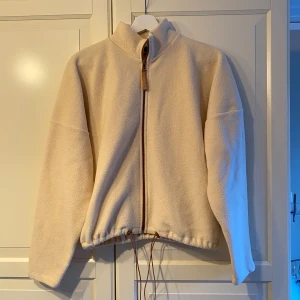 Beige fleecejacka med dragkedja - Mysig beige fleecejacka perfekt för kyliga dagar. Den har en dragkedja framtill och justerbar dragsko i nederkant för bästa passform. Jackan är långärmad och har en hög krage som håller dig varm. Perfekt för höst och vår!