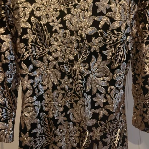 Svart klänning med guldblommor - Snygg svart klänning från ADM med glittriga guldblommor över hela tyget. Den har en djup v-ringning och trekvartsärmar, perfekt för festliga tillfällen. Klänningen är i mycket bra skick och ger en lyxig känsla med sitt blommiga mönster.