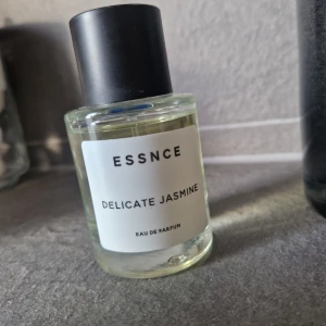 Essnce Delicate Jasmine Eau de Parfum - Säljer en elegant flaska med Essnce Delicate Jasmine Eau de Parfum. Doften är fräsch och blommig med en tydlig jasminnot. Flaskan är stilren och genomskinlig med en svart kork, perfekt för den som älskar en subtil och sofistikerad doft.