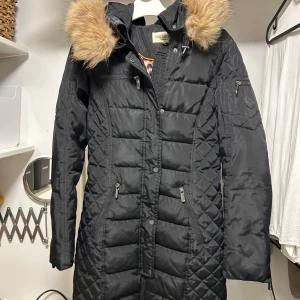 Svart vinterjacka med pälsdetaljer - Säljer en snygg svart vinterjacka från Rocco Baroco. Den har en mysig pälsfodrad huva och praktiska fickor med dragkedjor. Perfekt för kalla vinterdagar! Jackan är i fint skick och har en figursydd passform. 