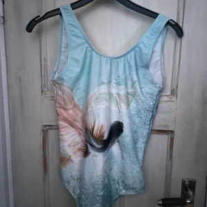 Ny mönstrad baddräkt från Molo - Superfin baddräkt från Molo med ett vackert fiskmotiv i mjuka pastellfärger. Den har en öppen rygg. Materialet är av hög kvalitet och erbjuder UV-skydd på 40+. Helt ny med taggar kvar! Butiksägaren har klippt svarta lappen i två bitar.