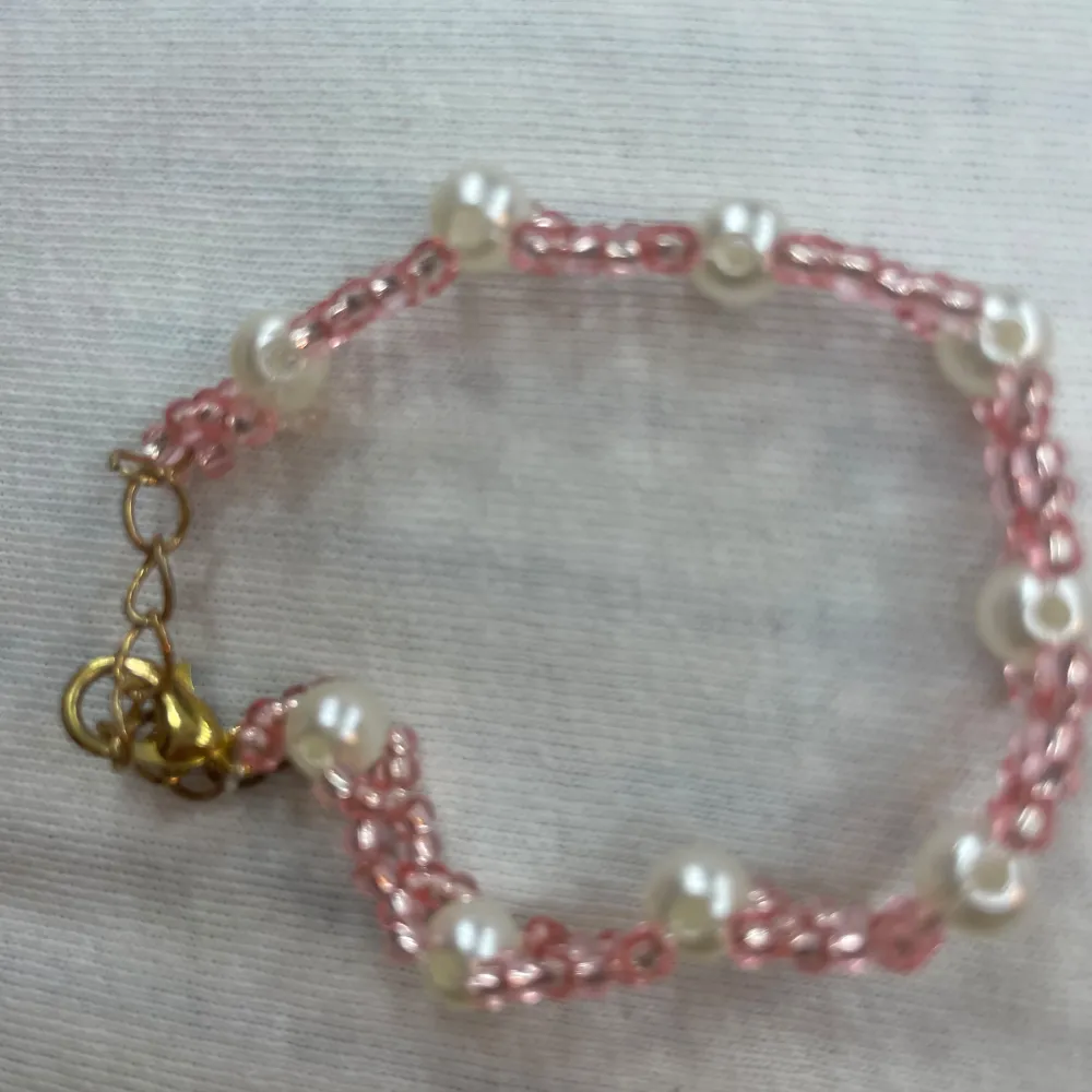 Ett superfint armband med rosa flätad design och vita pärlor. Perfekt för att ge en söt och elegant touch till din outfit. Armbandet har en guldfärgad låsning som gör det enkelt att ta på och av. Passar perfekt till både vardag och fest!. Asusteet.