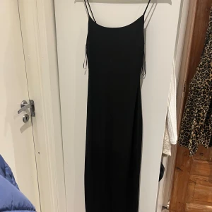 Svart klänning - Elegant svart långklänning med tunna axelband, perfekt för festliga tillfällen. Klänningen har en stilren och tidlös design som passar både till nyår och andra högtider. Materialet ser ut att vara mjukt och följsamt, vilket ger en bekväm passform. Endast använd en gång