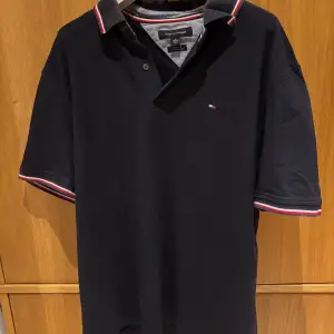 Säljer en klassisk mörkblå pikétröja från Tommy Hilfiger i storlek XL. Tröjan har korta ärmar och en snygg krage med röd och vit rand. Perfekt för både vardag och festliga tillfällen. Den är i mycket bra skick och redo för en ny ägare!