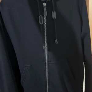 Svart huvtröja från Burberry - Snygg svart huvtröja från Burberry med dragkedja och klassiskt rutigt mönster på insidan av huvan. Perfekt för en stilren look. Den är i stl S men den är stor i storlek.