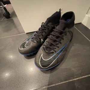 Nike fotbollsskor med blå sula - Snygga svarta Nike fotbollsskor med blå sula och vit swoosh. Skorna har snörning och är perfekta för fotbollsspel på olika underlag. De har en modern design med detaljer i blått och vitt. men är lite sönder i baksidan. Och de är gruskor. Nya kostar 1200kr men jag säljer för 400kr.
