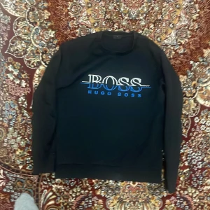 Svart tröja från Hugo Boss - Snygg svart tröja från Hugo Boss med blått och vitt tryck på bröstet. Perfekt för en stilren look. Tröjan har långa ärmar och rund halsringning.