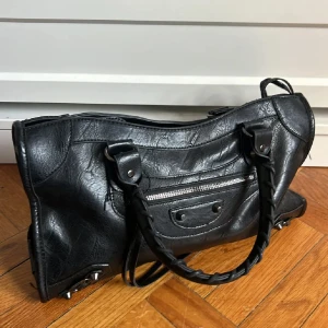 Trendig väska - Snygg svart skinnväska med flätade handtag och avtagbar axelrem. Väskan har en dragkedja framtill och metallfötter på undersidan för extra skydd. Perfekt för att bära dina dagliga nödvändigheter med stil. Bra skick! 