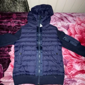 Mörkblå dunjacka från Moncler - Säljer en snygg mörkblå dunjacka från Moncler med huva och dragkedja. Jackan har ribbade muddar och en praktisk ficka på ärmen. Perfekt för kyliga dagar.