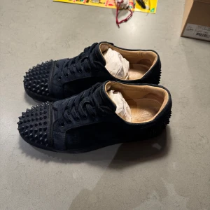 Mörkblåa sneakers med nitar (Louboutin) - Mörkblåa sneakers från Louboutin med nitar på tå och häl. Skorna har en stilren design med snörning och är tillverkade i mocka. Perfekta för att ge din outfit en edgy touch.   Orginalkartong, extra vita snören, kvitto mm