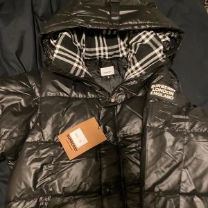 Svart dunjacka från Burberry S/M  - Snygg svart dunjacka från Burberry med huva och klassiskt rutigt foder. Jackan har dragkedja och tryckknappar framtill samt Burberry London England-logga på bröstet. Perfekt för kyliga dagar. Pris kan diskuteras. Storleken fungerar på både S och M kan utöver byta jacka 