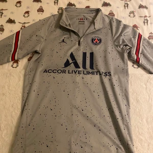 Grå PSG fotbollströja med Jordan-märke - Säljer en grå PSG fotbollströja med Jordan-logga och texten 'Accor Live Limitless' på framsidan. Tröjan har långa ärmar med röda och svarta ränder samt texten 'Visit Rwanda' på baksidan. Perfekt för fotbollsfans! Och säljs för bara 450kr nöstan aldrig använd topp skick hör av er via mer frågor 