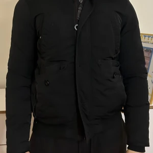 Svart dunjacka från Tom Tailor - Snygg svart dunjacka från Tom Tailor med huva och pälsdetalj. Jackan har dragkedja och knappar framtill samt praktiska fickor. Perfekt för kyliga dagar. Jackan är även i perfekt skick, pris kan diskuteras 