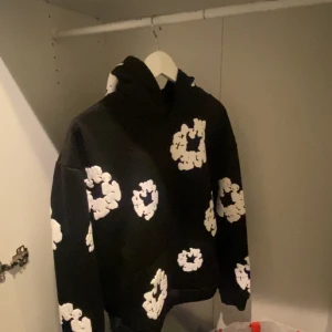 Svart hoodie med blommönster från Denim Tears - Snygg svart hoodie från Denim Tears med vita blommönster. Perfekt för en avslappnad stil med en unik touch. Hoodien har en klassisk passform och är tillverkad i ett mjukt material. Har även den digitala kvittot