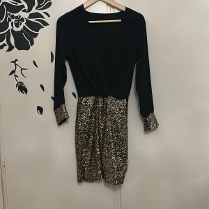 Svart och guldglittrig nyårs klänning - Snygg klänning med svart överdel och guldglittrig kjol. Långärmad design med glittriga detaljer vid ärmslut. Perfekt för en festlig kväll!