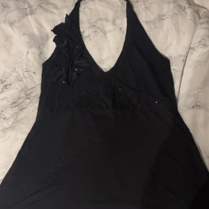 Svart topp från Gina Tricot - Snygg svart topp från Gina Tricot med v-ringning och dekorativa blommor på ena axeln. Klänningen har paljettdetaljer som ger en glittrig effekt. Perfekt för en kväll ute!
