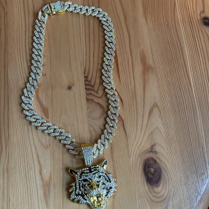 Guldhalsband med tigerhänge - Snyggt guldhalsband med ett stort tigerhänge täckt av glittrande stenar. Kedjan är kraftig och har en säker låsmekanism. Perfekt för att göra ett statement.