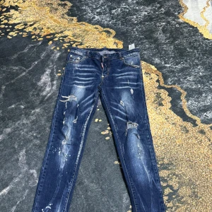 Blå jeans med slitningar - Säljer ett par blå jeans med coola slitningar och detaljer. Perfekta för en avslappnad stil. De har en klassisk femficksdesign och knappgylf. Köpt för 900 kr 