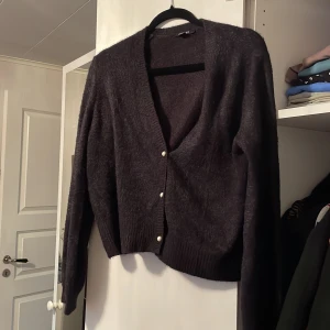 Mörkgrå kofta med pärlknappar - Säljer en elegant mörkgrå kofta med v-ringning och pärlknappar. Koftan har långa ärmar och är tillverkad i ett mjukt material som ger en lyxig känsla. Perfekt för kyliga dagar!