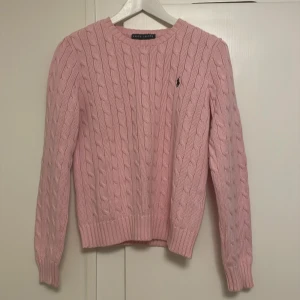 Rosa kabelstickad tröja från Ralph Lauren - Säljer en snygg rosa kabelstickad tröja från Ralph Lauren. Tröjan har en klassisk design med rund hals och det ikoniska logotypbroderiet på bröstet. Perfekt för en stilren och bekväm look.