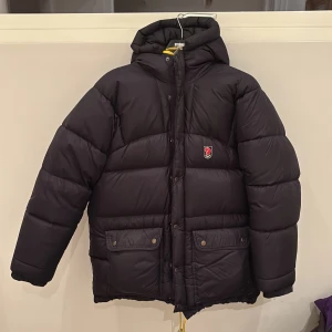 Svart Fjällräven expedition dunjacka - Säljer en svart dunjacka från Fjällräven Polar med huva och knäppning fram. Jackan har två stora fickor med knappar och en innerficka med dragkedja. Perfekt för kalla vinterdagar.  Modell är expedition. Använd men i bra skick.