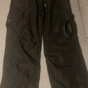 Svarta cargopants med fickor i polyester storlek L - Snygga svarta cargopants med praktiska fickor på sidorna. Perfekta för en avslappnad och funktionell stil. Byxorna har en loose passform och är tillverkade i ett slitstarkt material. Midjan har stretch funktion och storleken är Large och är tillverkade i polyester.