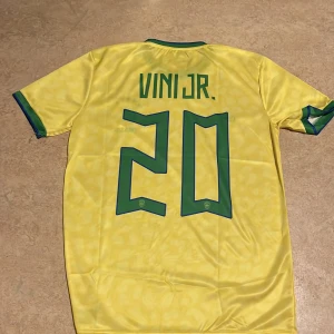 Gul Brasilien fotbollströja  - Säljer en gul Brasilien fotbollströja från Nike med korta ärmar och gröna detaljer. Tröjan har spelarnamnet 'VINI JR.' och nummer 20 på ryggen. Perfekt för fotbollsfans!